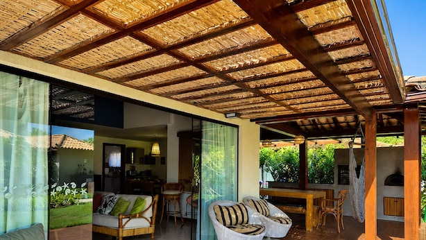 A modern outdoor living area with a wooden ceiling, glass walls, and wicker furniture. - Villa Serena | Vacation Home for Families and Groups (Armação dos Búzios)