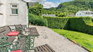 Eine Terrasse mit Tisch und Bänken, ein weißes Gebäude mit einem Fenster und ein grüner Heckenstreifen.