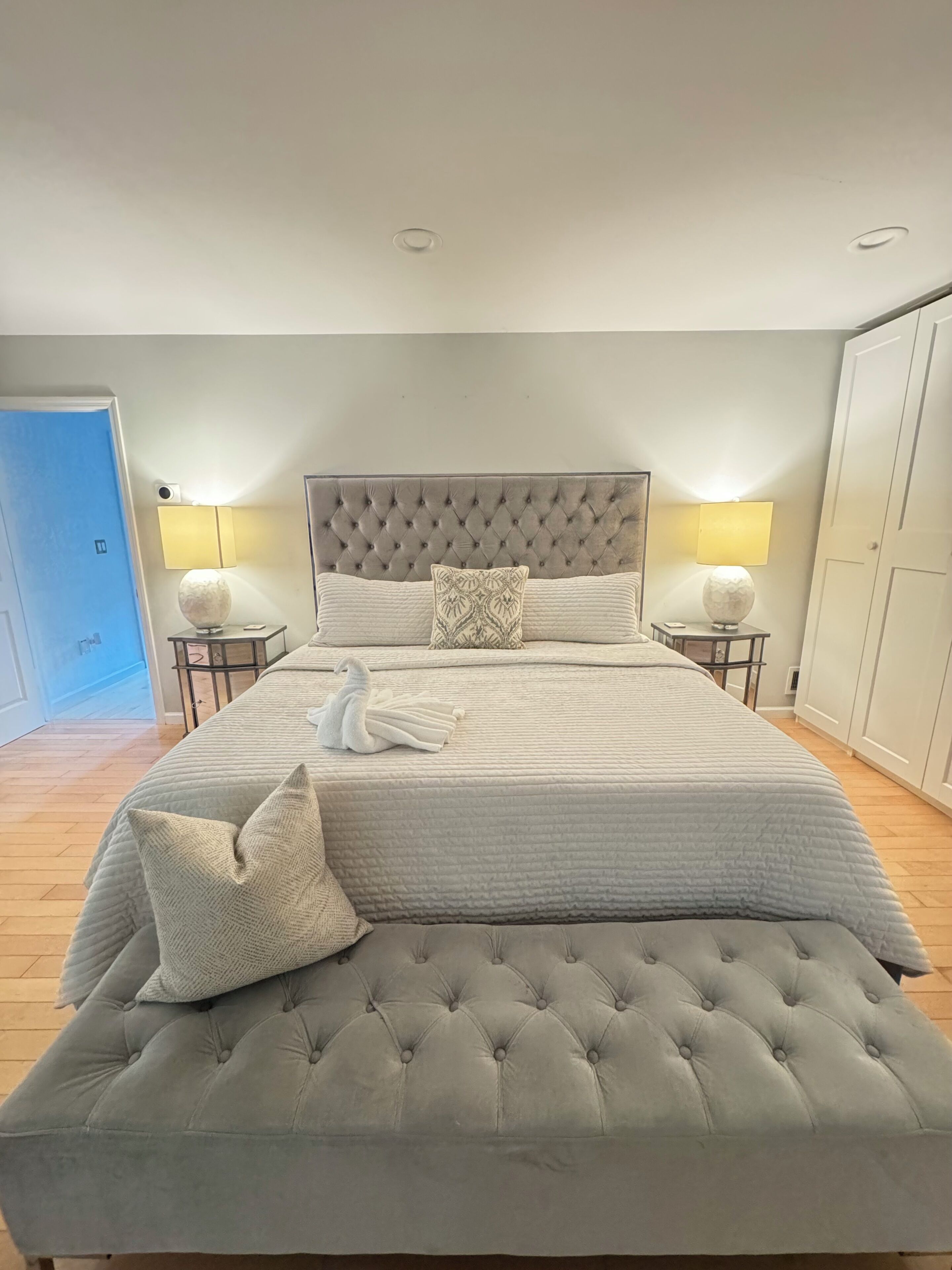 Een net opgemaakt bed met een gestoffeerd hoofdbord, twee bedlampjes en een handdoek in de vorm van een zwaan.