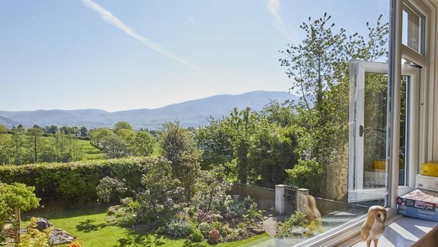 A view from a window overlooking a garden with a variety of plants and a distant mountain range. - 2 Bed in Cockermouth (oc-sz381) (Wythop)