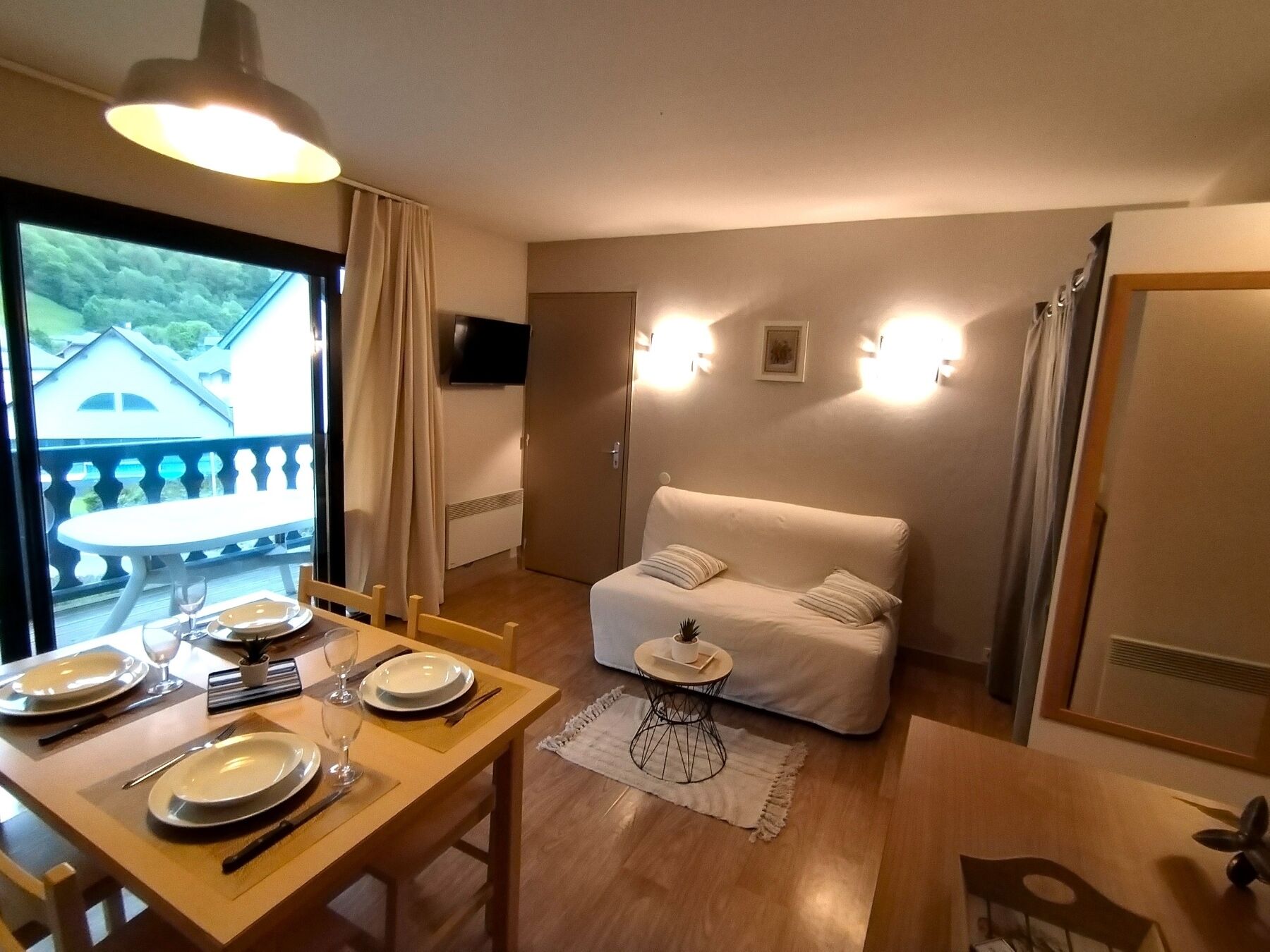 Une pièce comprenant une table à manger dressée pour deux personnes, un canapé, une petite table d’appoint et un téléviseur fixé au mur.