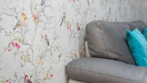 A grey sofa with a teal pillow, a floral wallpaper with birds, and a white wall. - 2 Bed in Keswick (oc-sz215) (Keswick)