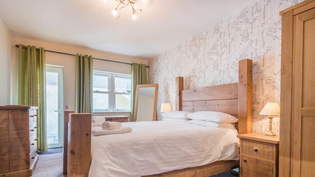 A bedroom with a wooden bed, a nightstand with a lamp, a dresser, and a mirror. - 2 Bed in Keswick (oc-sz215) (Keswick)