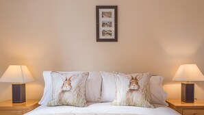 Uma cama bem arrumada com cobertura acolchoada, dois travesseiros com desenho de coelho, duas luminárias de cabeceira e um quadro na parede.