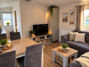 Ein modernes Wohnzimmer mit Flachbildfernseher, grauen Sofas, einem Holzesstisch und einer Küchenzeile im Hintergrund.