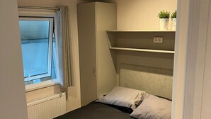 Un dormitorio con cama, radiador, ventana con cortinas, armario y estante con plantas.