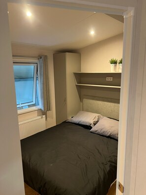 A bedroom with a bed, a radiator, a window with curtains, a wardrobe, and a shelf with plants. - Chalet 'Duinzicht' with Shared Pool, Private Garden and Wi-Fi (Kamperland)