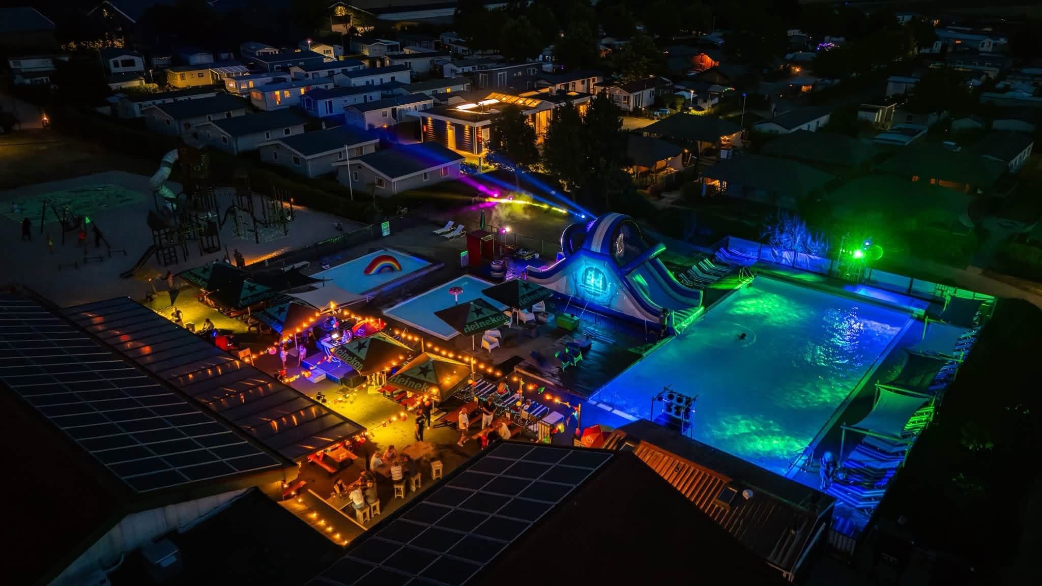 Un espace extérieur avec une piscine et un toboggan, entouré de chaises longues et de parasols, ainsi qu’une scène équipée d’un système d’éclairage.