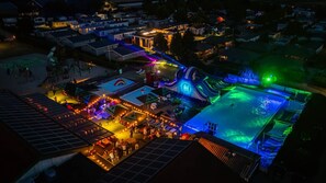 Un área de piscina al aire libre con tobogán, rodeada de sillas de descanso y sombrillas, y un escenario con iluminación.