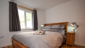 Un dormitorio con cama de madera, una mesita de noche con lámpara, una ventana con cortinas y una bandeja con tetera y tazas.