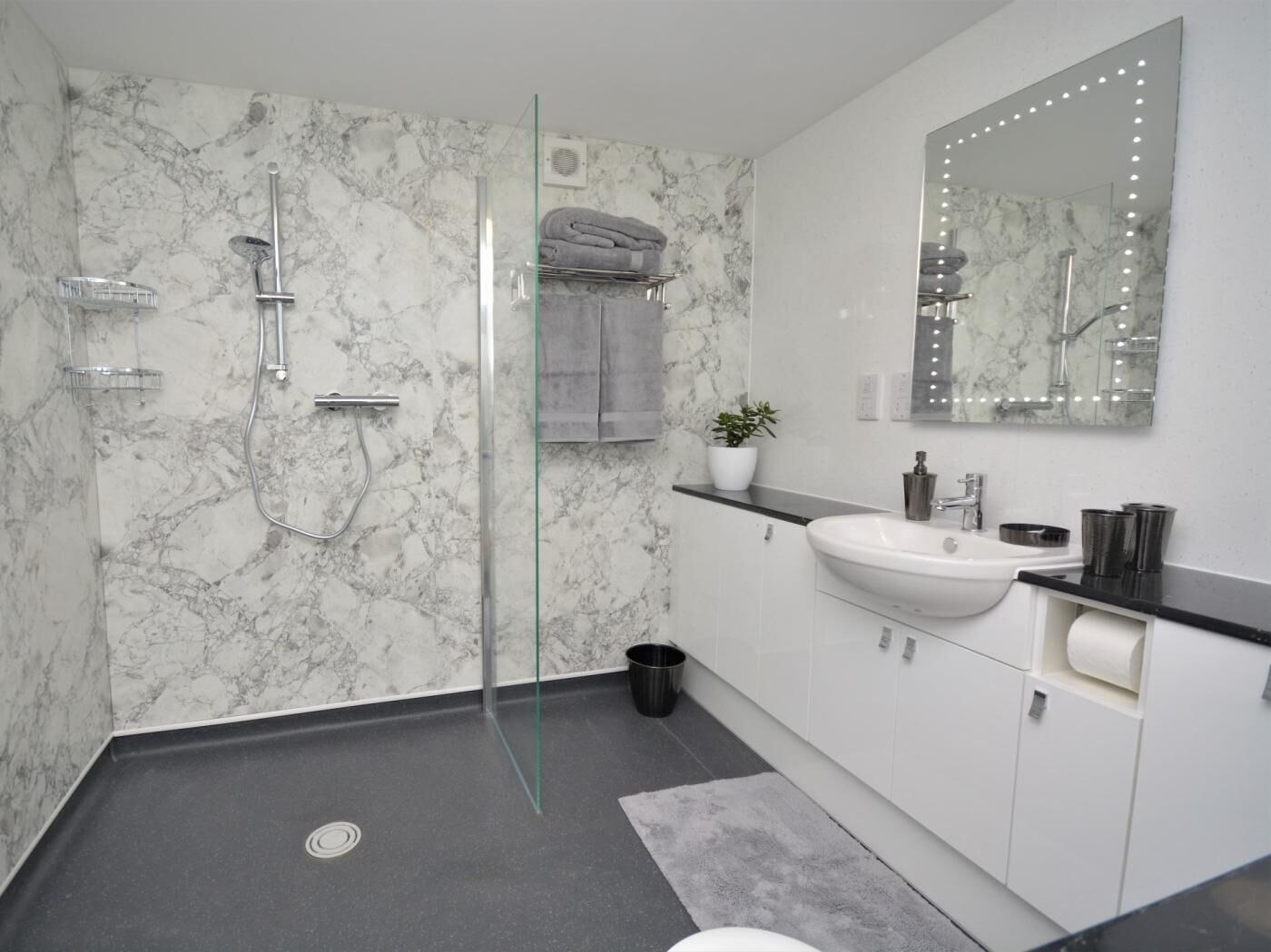 A modern bathroom with a glass shower enclosure, a marble wall, a sink with a vanity, and a mirror with lights.