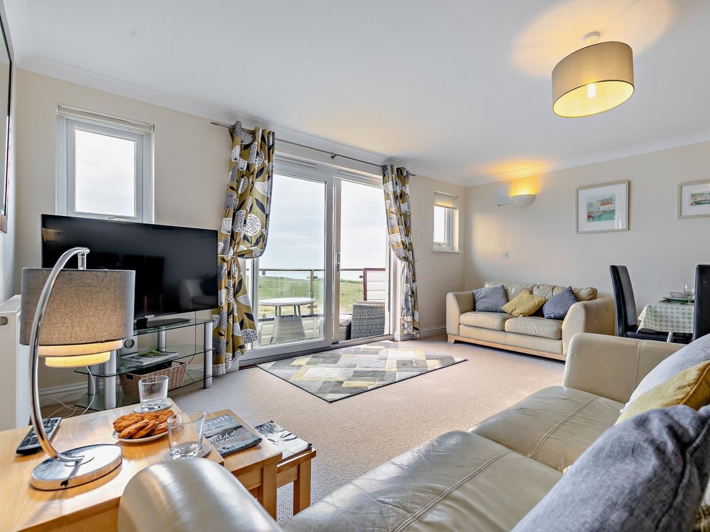A modern living room with a flat-screen TV, a sofa, a dining table with a lamp, and a view through large windows.