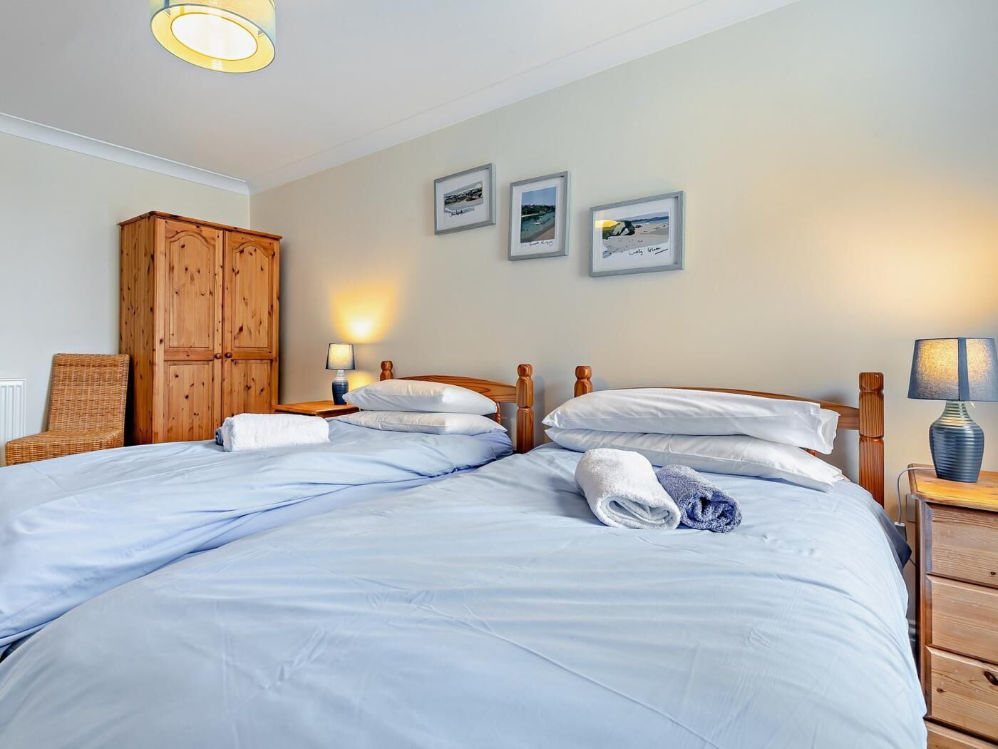 A double bed with white linens, a wooden wardrobe, a wicker chair, and three framed pictures on the wall.