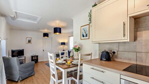 Una cocina moderna con armarios blancos, encimera de madera y horno empotrado. Hay una zona de comedor con una mesa preparada para dos personas, sillas blancas y un sofá gris.