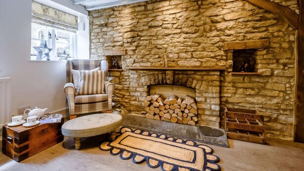 A cozy room with a stone fireplace, a plaid armchair, a round ottoman, and a wooden chest of drawers. - 2 Bed in Cheltenham (oc-46677) (Bourton-on-the-Water)