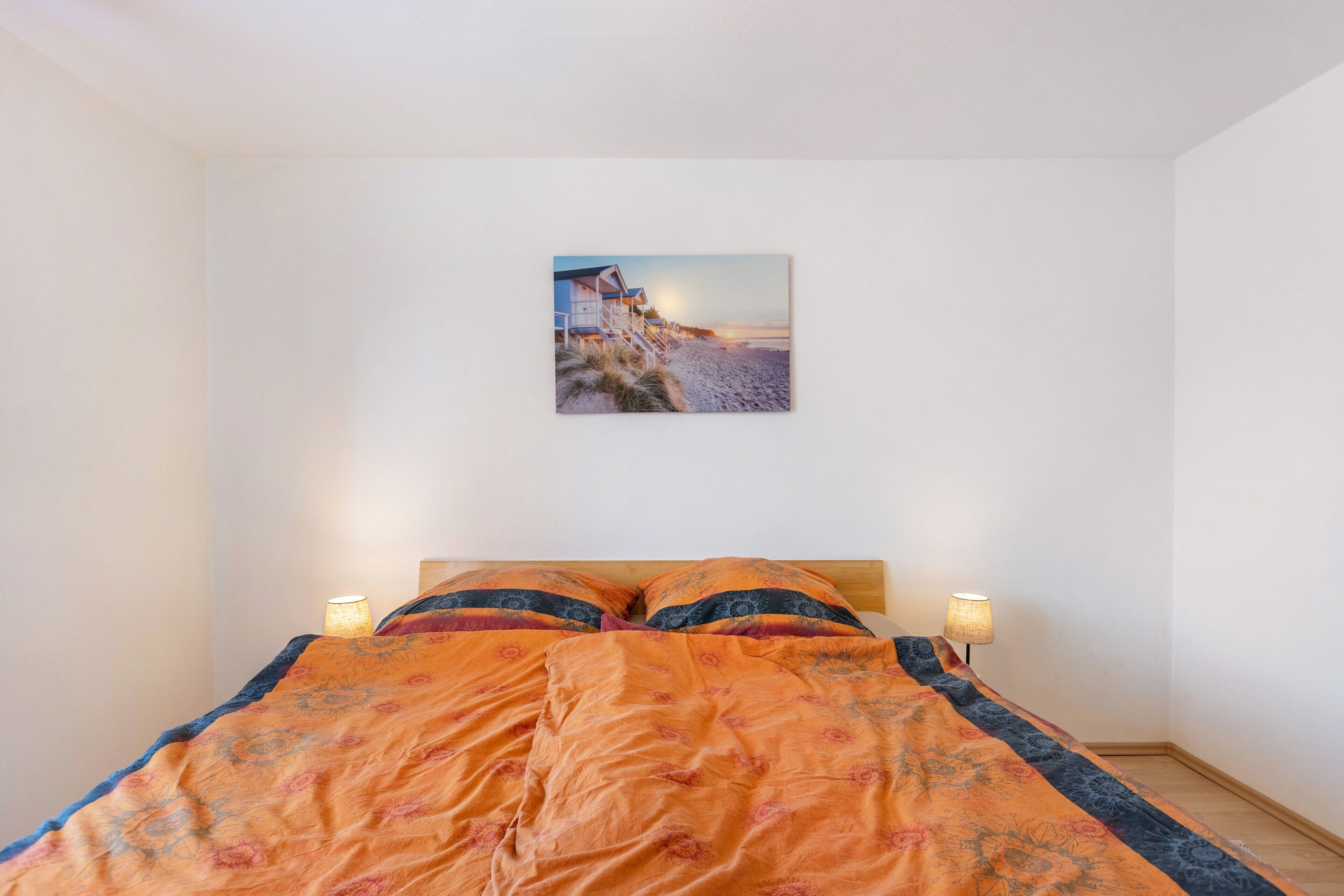 A bed with an orange floral bedspread, two bedside lamps, and a beach scene painting on the wall.