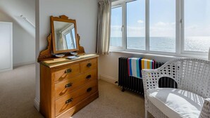 A room with a wooden dresser, a mirror, a wicker chair, a radiator, and a window with a view of the sea. - 3 Bed in Bexhill On Sea (oc-bt037) (Bexhill-on-Sea)