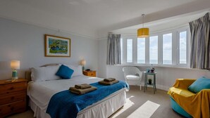 A bedroom with a bed, two bedside tables, a chair, a small table, and a window with curtains. - 3 Bed in Bexhill On Sea (oc-bt037) (Bexhill-on-Sea)
