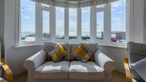 A room with a curved bay window, a beige sofa with patterned cushions, and a view of the sea. - 3 Bed in Bexhill On Sea (oc-bt037) (Bexhill-on-Sea)