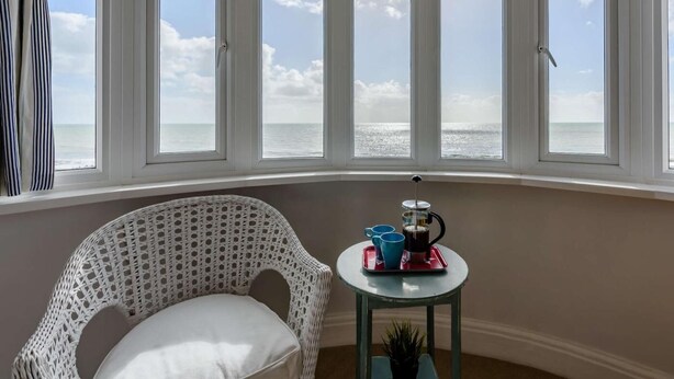 A room with a bay window overlooking the sea, a wicker chair, and a small table with a tray holding a coffee pot and cups. - 3 Bed in Bexhill On Sea (oc-bt037) (Bexhill-on-Sea)