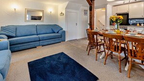 A living room with a blue sofa, a dining table with wooden chairs, a kitchen with white cabinets, and a black rug. - 4 Bed in Bideford (oc-46840) (Pusehill)