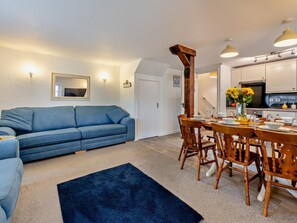 A living room with a blue sofa, a dining table with wooden chairs, a kitchen with white cabinets, and a black rug. - 4 Bed in Bideford (oc-46840) (Pusehill)