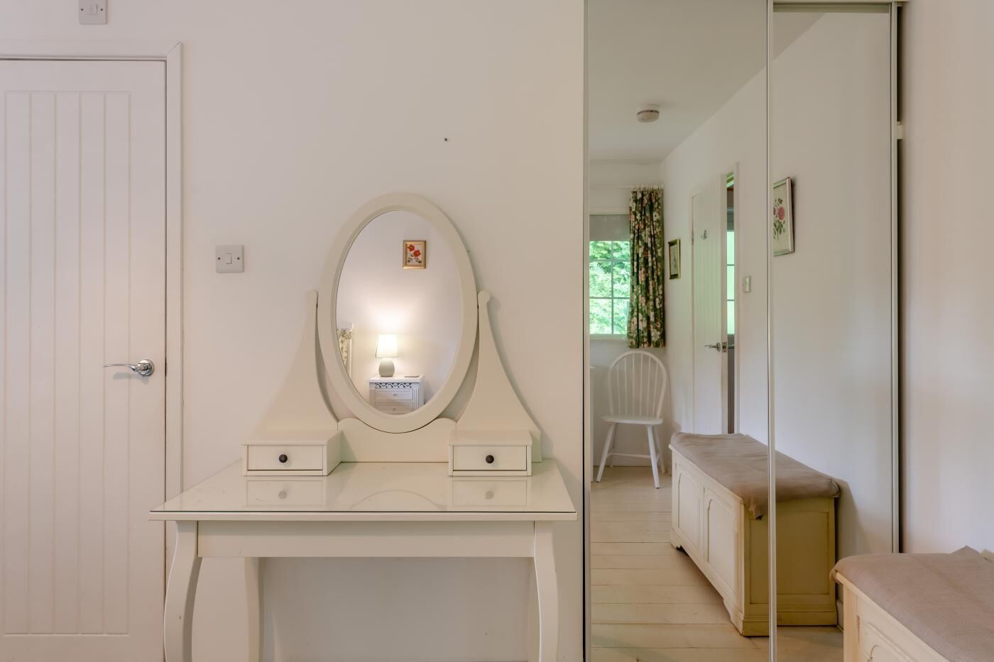 Ein Zimmer mit einem weißen Schminktisch, einem Spiegel, zwei Schubladen, einem kleinen Tisch mit Lampe und einem weißen Stuhl.