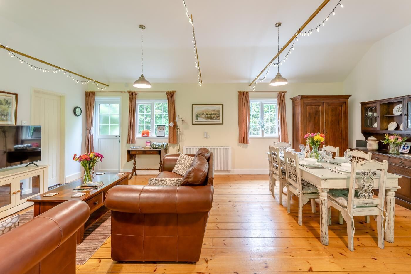 A spacious living room with a brown leather sofa, wooden dining table, and chairs. There is a television on a stand, a fireplace, and a window with curtains.