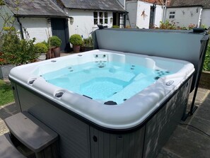 A hot tub with a built-in seat and steps, surrounded by a garden with potted plants and a house in the background. - 5 Bed in Hurst Green (oc-46916) (Robertsbridge)