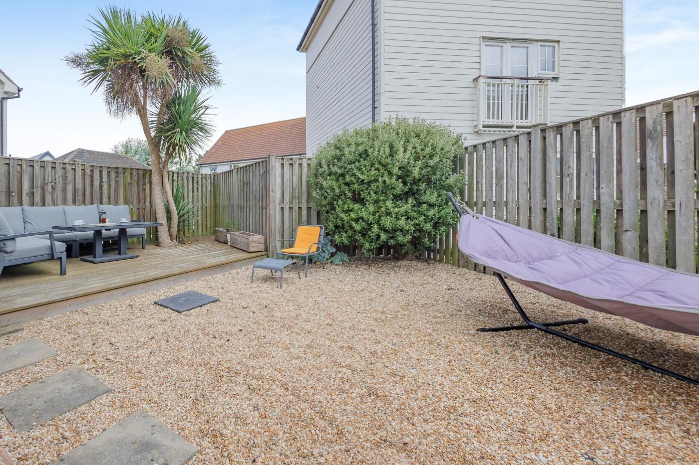 Ein Garten mit einer Holzterrasse, einem Patio-Tisch, einer Couch, einem Stuhl, einem kleinen Busch und einer Hängematte.