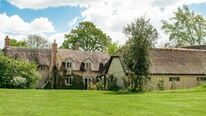 A traditional stone house with a thatched roof, a grassy lawn, and a large tree in the background. - 5 Bed in Sturminster Newton (oc-76458) (Woodrow)