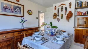 Uma sala de jantar com uma mesa posta para quatro pessoas, um candelabro, um relógio e uma estante de livros.