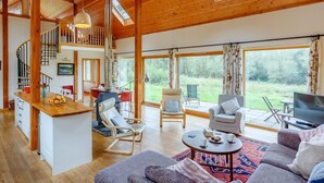 A spacious living area with a wooden ceiling, a kitchen with a central island, a dining area, and a large window offering a view of the outdoors. - 4 Bed in Fort William (oc-78875) (Muirshearlich)