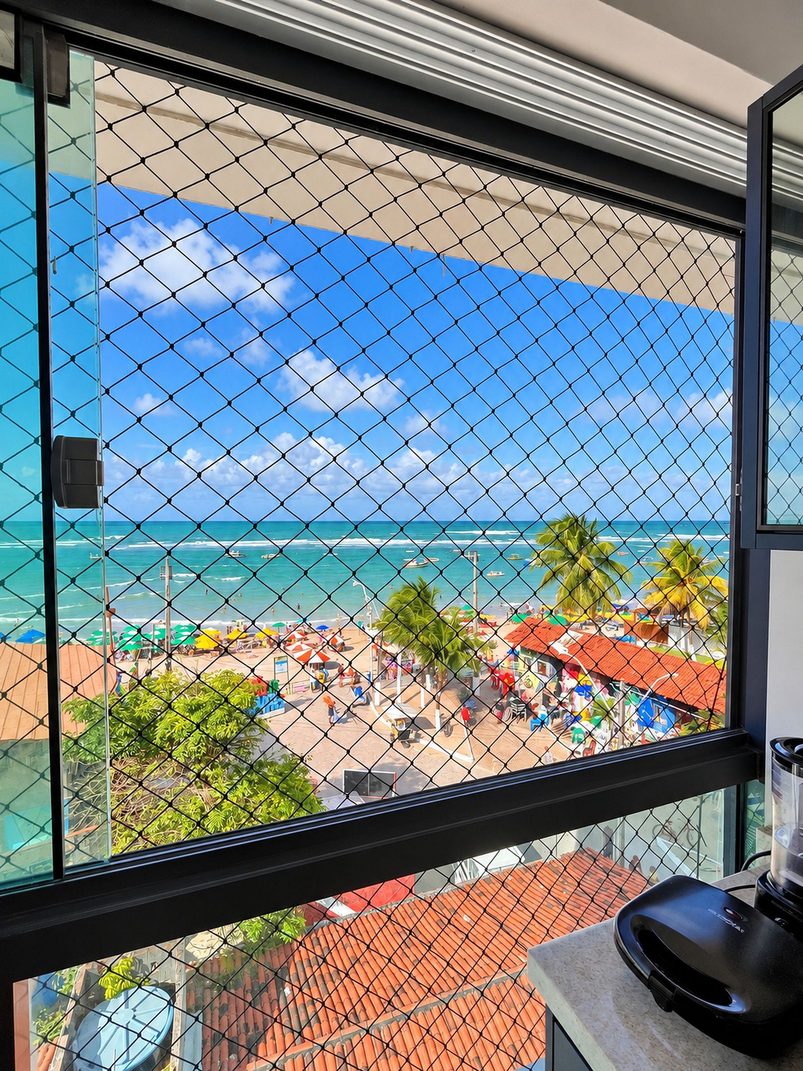Una habitación con vista a una playa, visible a través de una ventana con una cerca de alambre.