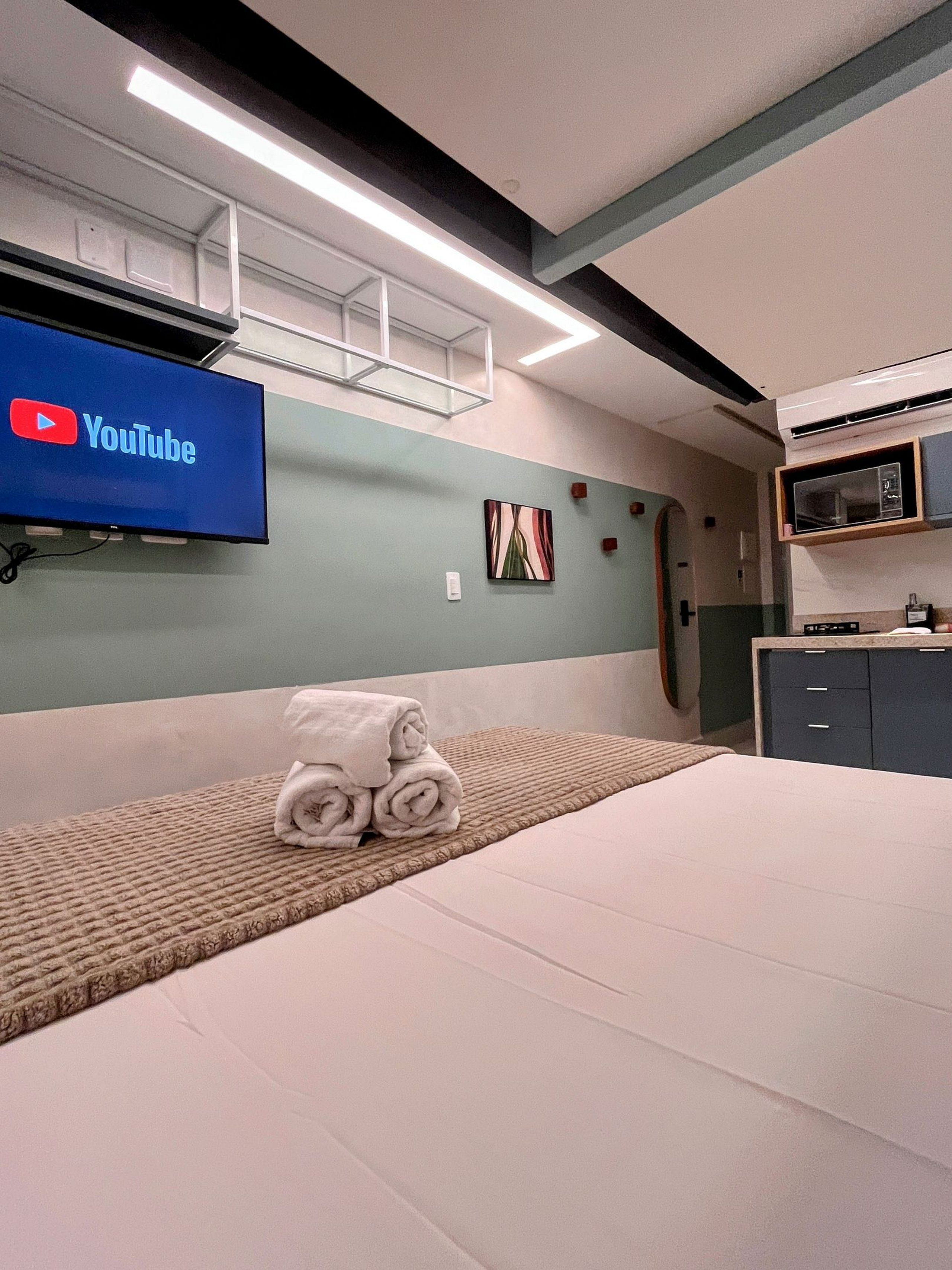 Une chambre d’hôtel avec un lit, un téléviseur à écran plat fixé au mur, une pile de serviettes, un petit tableau et un four à micro-ondes dans le coin cuisine.