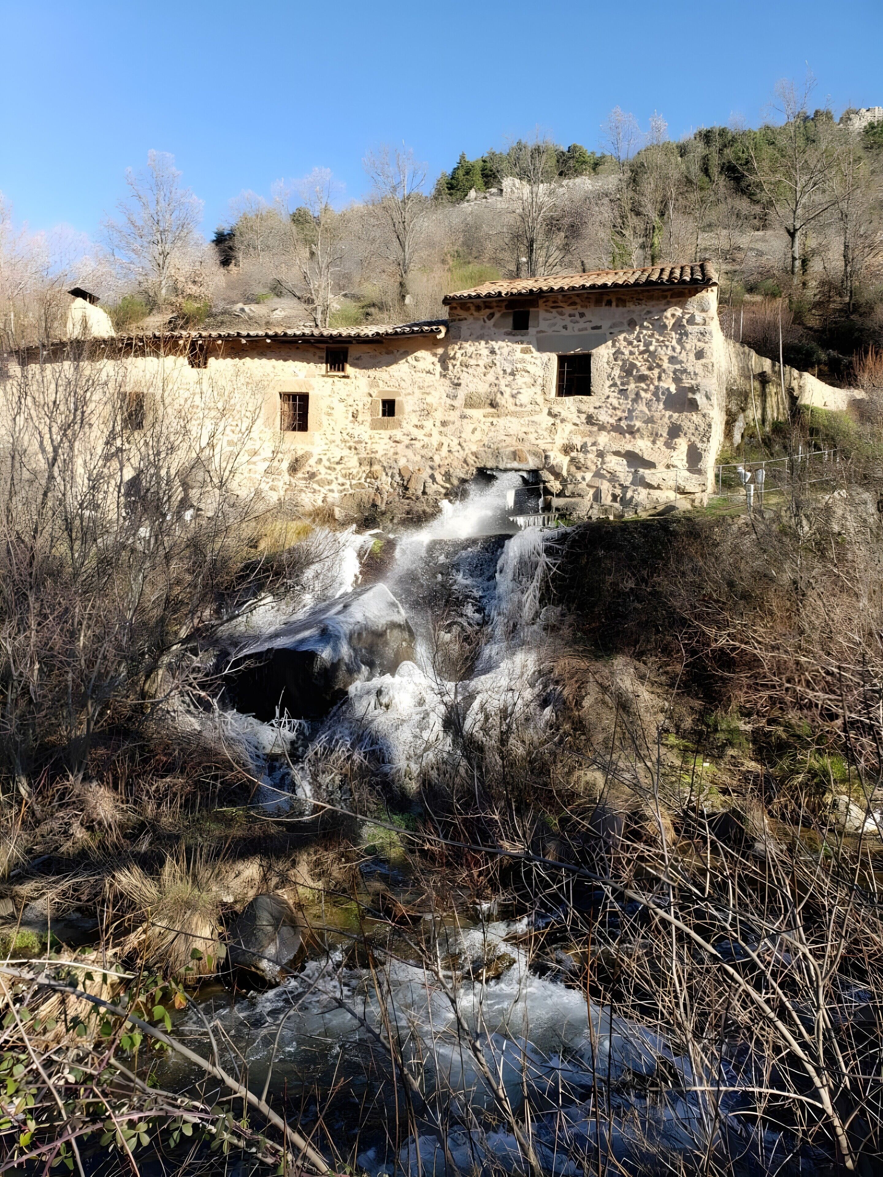 A rustic stone building with a small waterfall, surrounded by dry vegetation and trees.