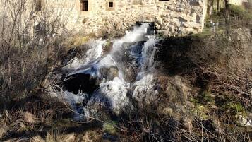 A rustic stone building with a small waterfall, surrounded by dry vegetation and trees.