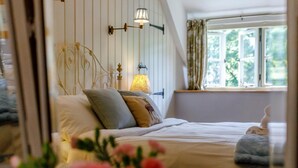 Ein Schlafzimmer mit einem Bett, Kissen, einer Nachttischlampe, einem Fenster mit Vorhängen und Blick ins Grüne.