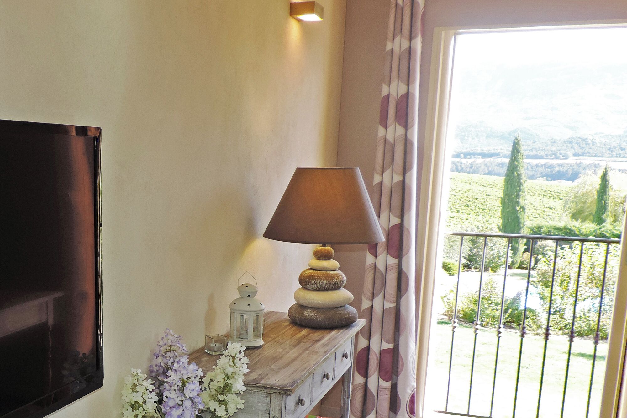 A room with a TV mounted on the wall, a wooden side table with a lamp, and a view of a vineyard through a balcony door.