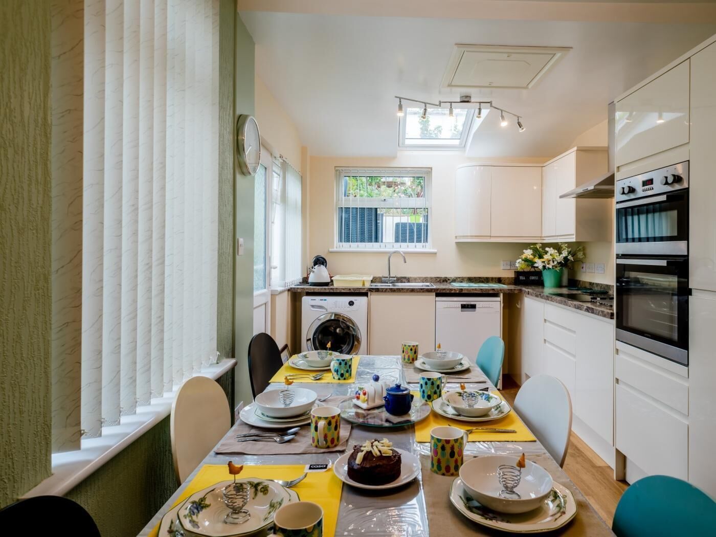 A modern kitchen with a dining area, featuring a built-in oven, a washing machine, and a window with blinds.