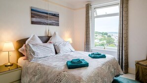 Un lit bien fait, avec une couvre-lit à motifs, deux oreillers et deux serviettes pliées. On y trouve une lampe de chevet, un tableau au mur et une fenêtre avec des rideaux.