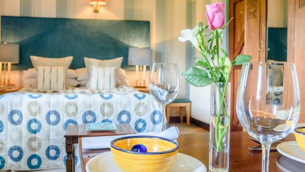 Ein Hotelzimmer mit Bett, Nachttischen, einer Vase mit Blumen und einem Esstisch mit Schüssel und Weingläsern.