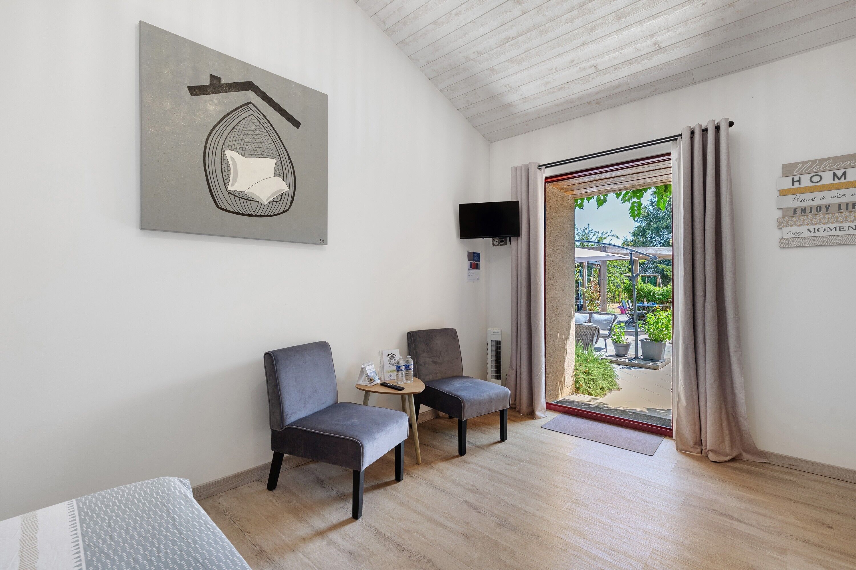A room with two chairs, a small table with a remote control, a flat-screen TV mounted on the wall, and a door leading to an outdoor area with a view of greenery.