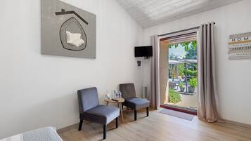 A room with two chairs, a small table with a remote control, a flat-screen TV mounted on the wall, and a door leading to an outdoor area with a view of greenery.