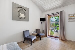 A room with two chairs, a small table with a remote control, a flat-screen TV mounted on the wall, and a door leading to an outdoor area with a view of greenery.