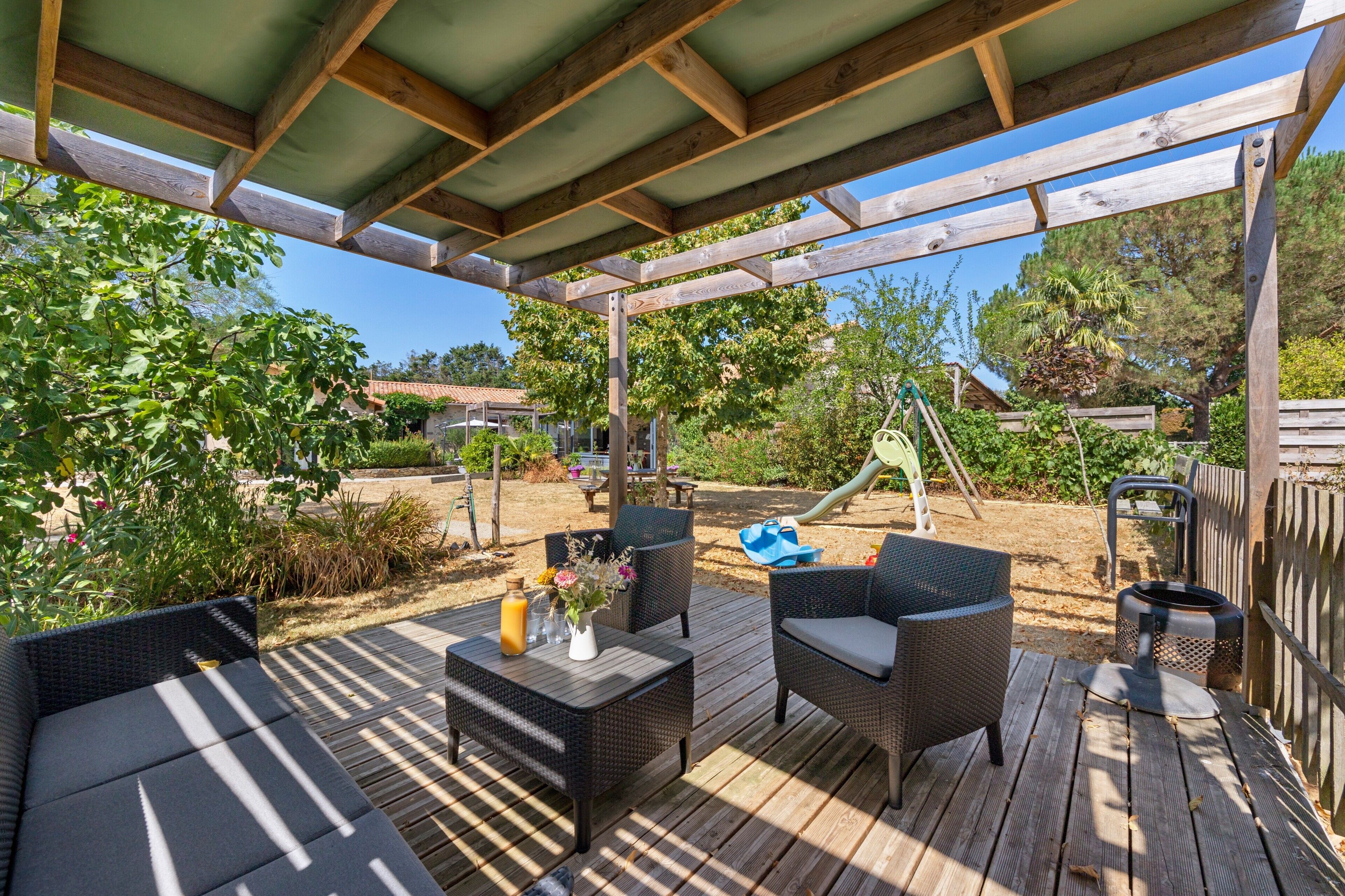 Une terrasse extérieure avec des meubles en osier, une table avec des boissons et, au fond, une aire de jeux.