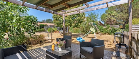 Een terras buiten met rieten meubilair, een tafel met drankjes en een speeltuin op de achtergrond.