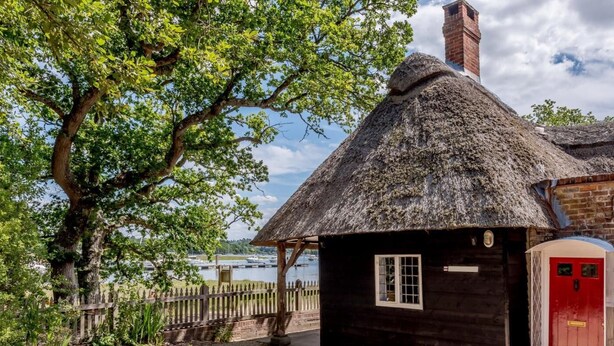 Een rieten huisje met een houten deur, een bakstenen schoorsteen en uitzicht op het water.
