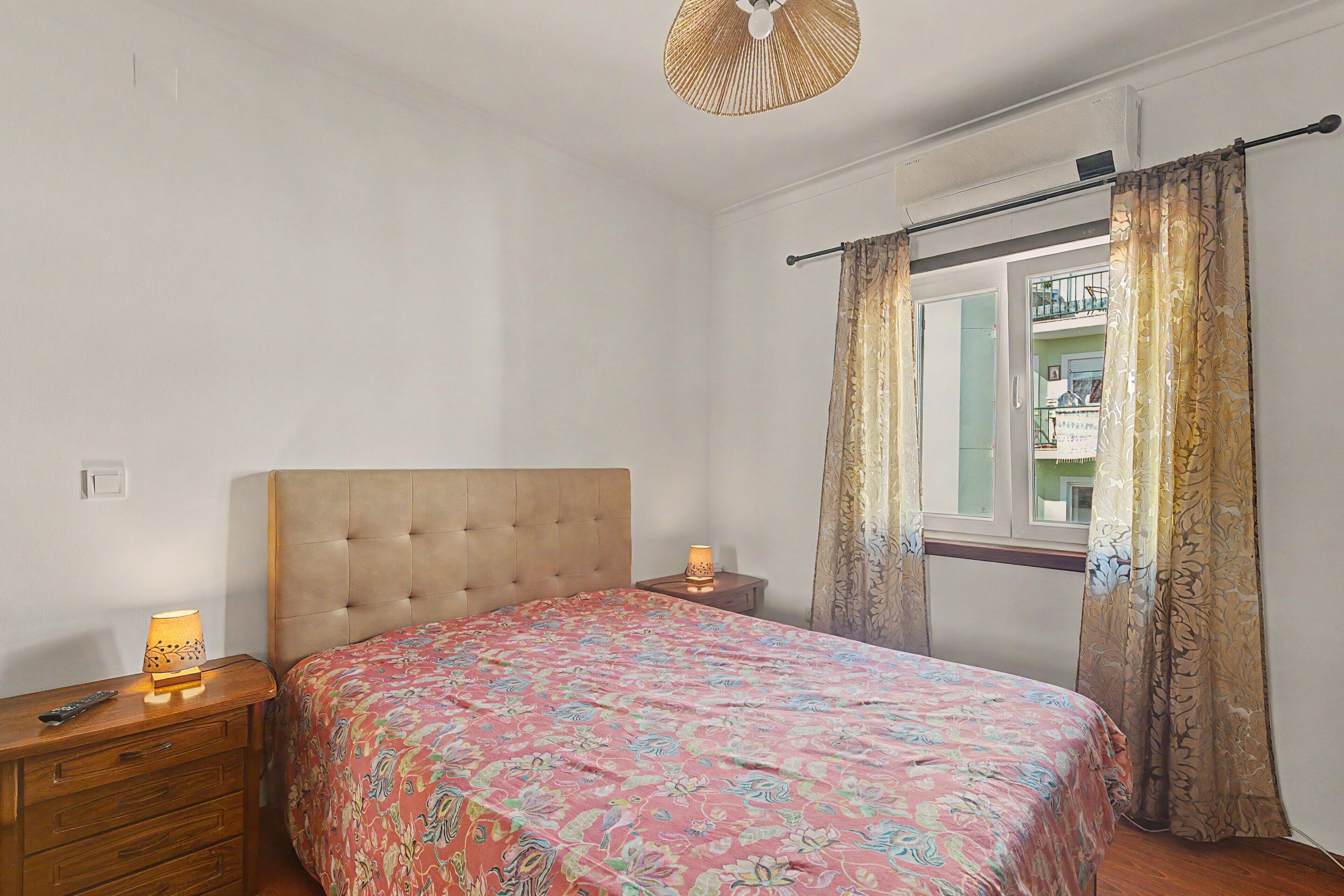 Un dormitorio con cama, lámparas de noche, ventana con cortinas, ventilador de techo y aire acondicionado instalado en la pared.