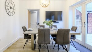Een eetgedeelte met een tafel, stoelen, een klok, een televisie en een deur naar een buitenruimte.